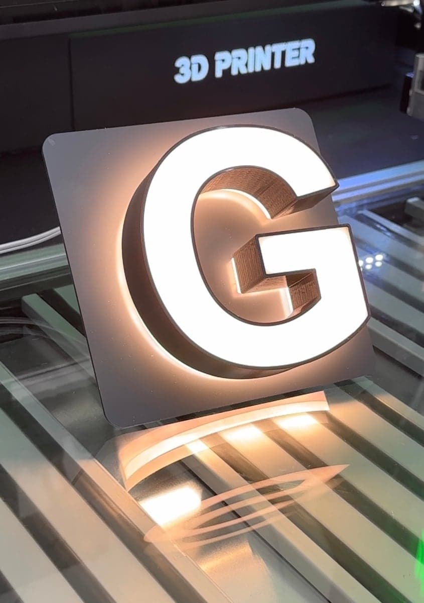 A white letter G standing on the printer bed, lit through its printed face and with a halo behind it